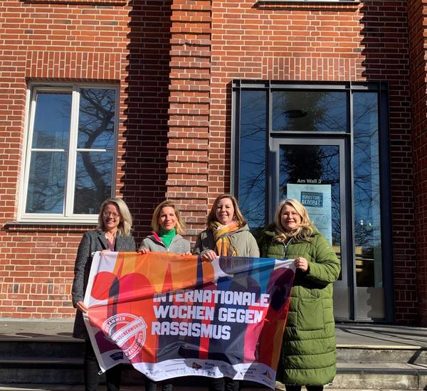 Dezernentin Anja Kerl, Maria Jonas von der Partnerschaft für Demokratie, Madlen Piertza aus der Integrationsarbeit des Landkreises Rostock und Ina-Maria Fahning, Leiterin des Sozialamtes Landkreis Rostock, präsentieren die Fahne vor dem Sitz der Kreisverwaltung in Güstrow.